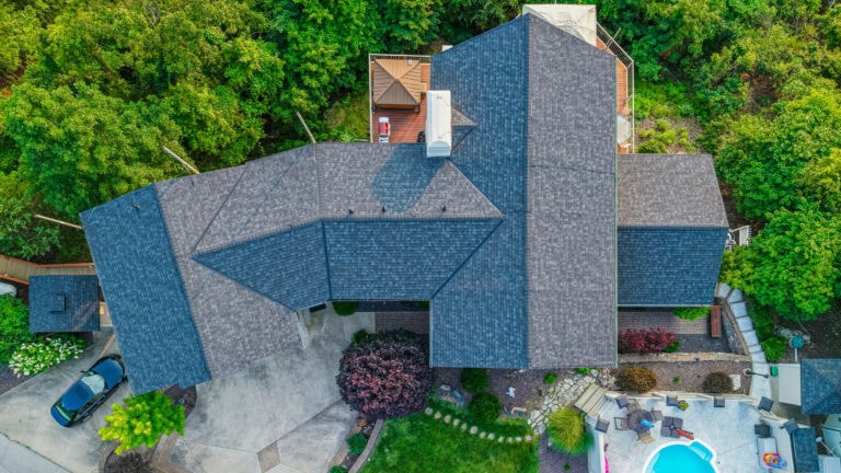 Aerial view of a residential roof with asphalt shingles surrounded by green trees, showcasing roof repair and installation services.